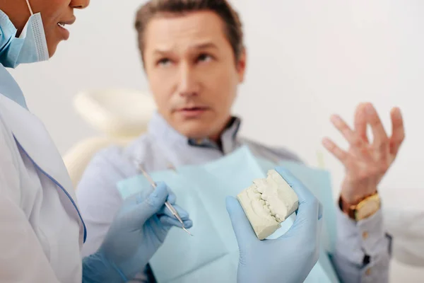 Селективный фокус африканского американского стоматолога в латексных перчатках с моделью зубов и зубным инструментом рядом с человеком — стоковое фото