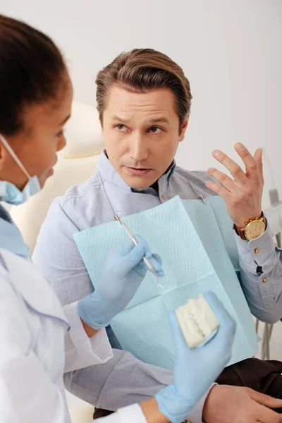 Foyer sélectif du dentiste afro-américain dans des gants en latex tenant le modèle de dents et l'instrument dentaire près du patient — Photo de stock
