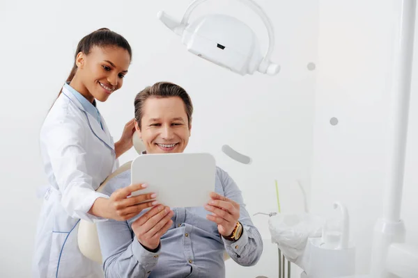 Zufriedene Patientin mit Zahnspange im Spiegel neben fröhlichem afrikanisch-amerikanischem Zahnarzt — Stockfoto