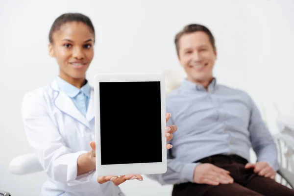 Messa a fuoco selettiva del paziente felice vicino allegro dentista afroamericano in possesso di tablet digitale con schermo bianco — Foto stock