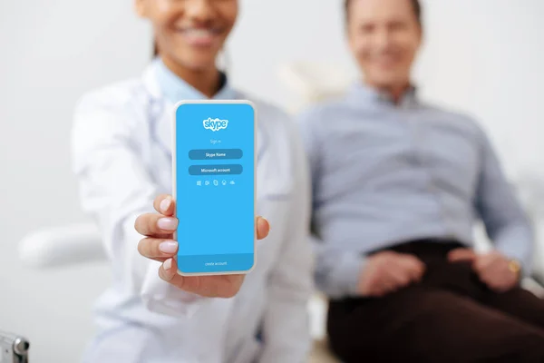 KYIV, UCRAINA - 12 MARZO 2020: focus selettivo di allegro dentista afroamericano che tiene smartphone con app skype sullo schermo vicino a paziente felice — Foto stock