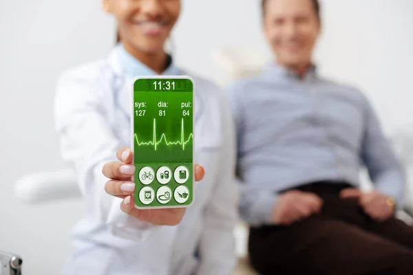 Foco seletivo de dentista americano africano alegre segurando smartphone com ritmo cardíaco na tela perto de paciente feliz — Fotografia de Stock
