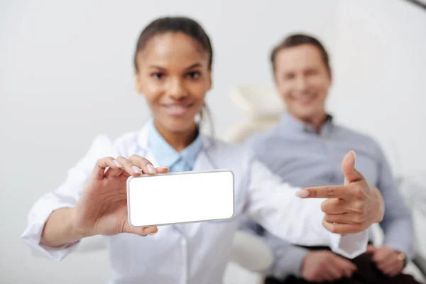 Foyer sélectif de gai dentiste afro-américain pointant du doigt smartphone avec écran blanc près patient heureux — Photo de stock