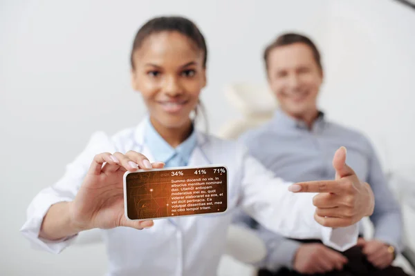 Selective focus of cheerful african american dentist pointing with finger at smartphone with medical app and lettering on screen near happy patient — Stock Photo