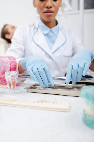 Vista ritagliata del dentista afroamericano in guanti di lattice toccare strumenti dentali inossidabili vicino al paziente — Foto stock