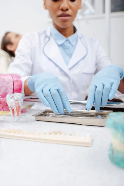 Visão recortada do dentista afro-americano em luvas de látex tocando instrumentos dentários inoxidáveis perto do paciente — Fotografia de Stock