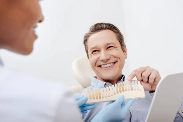 Избирательный фокус счастливого пациента, указывающего пальцем на палитру зубов рядом с веселым африканским американским стоматологом — стоковое фото