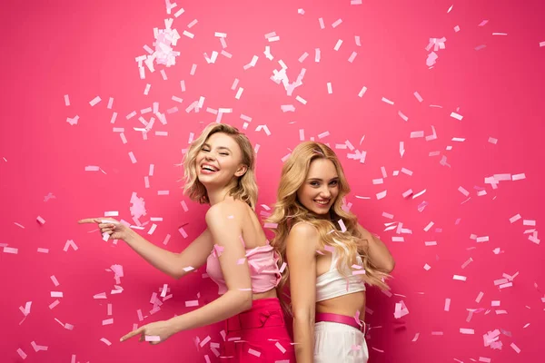 Side view of smiling blonde friends looking at camera and pointing with fingers under falling confetti on pink background — Stock Photo