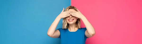 Panoramic shot of positive blonde woman covering eyes with hands on pink and blue background — Stock Photo