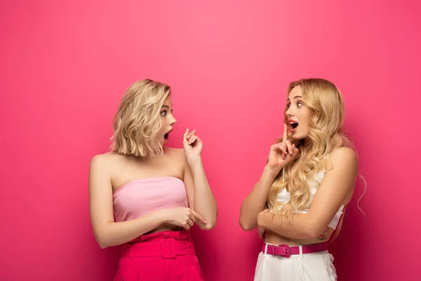 Chicas rubias impactadas mirándose y señalándose con los dedos sobre el fondo rosa - foto de stock