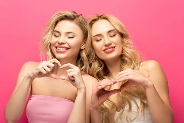 Sonrientes chicas rubias con los ojos cerrados mostrando el signo del corazón en el fondo rosa - foto de stock