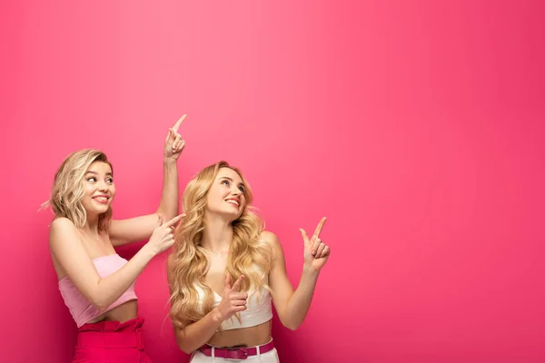 Chicas rubias felices señalando con los dedos y mirando hacia arriba en el fondo rosa - foto de stock