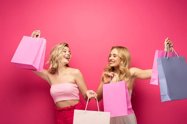 Happy blonde girls smiling while holding shopping bags on pink background — Stock Photo