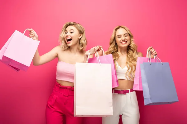 Alegre y sorprendida rubia amigos sosteniendo bolsas de compras sobre fondo rosa - foto de stock