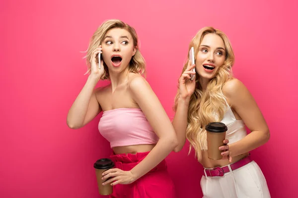 Aufgeregte blonde Freunde halten Pappbecher in die Höhe, während sie auf Smartphones auf rosa Hintergrund sprechen — Stockfoto