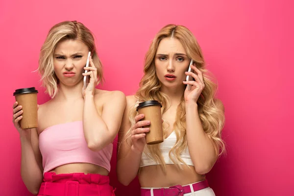 Verwirrte blonde Mädchen mit Pappbechern reden auf Smartphones auf rosa Hintergrund — Stockfoto
