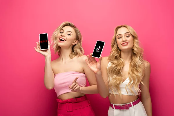 KYIV, UCRANIA - 10 de marzo de 2020: Amigos rubias positivos sosteniendo teléfonos inteligentes con aplicaciones de netflix y TikTok sobre fondo rosa - foto de stock