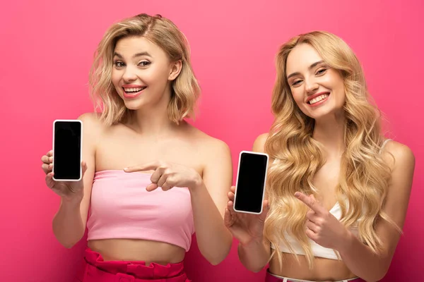Chicas rubias sonrientes apuntando con los dedos a teléfonos inteligentes con pantallas en blanco sobre fondo rosa - foto de stock