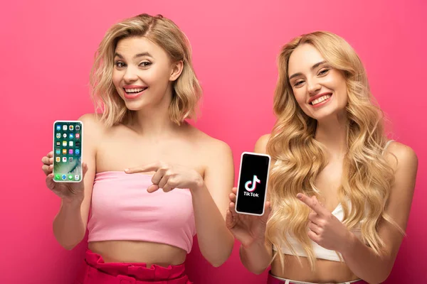 KYIV, UCRANIA - 10 de marzo de 2020: Amigos rubias sonrientes apuntando a teléfonos inteligentes con pantalla de iphone y aplicación TikTok sobre fondo rosa - foto de stock