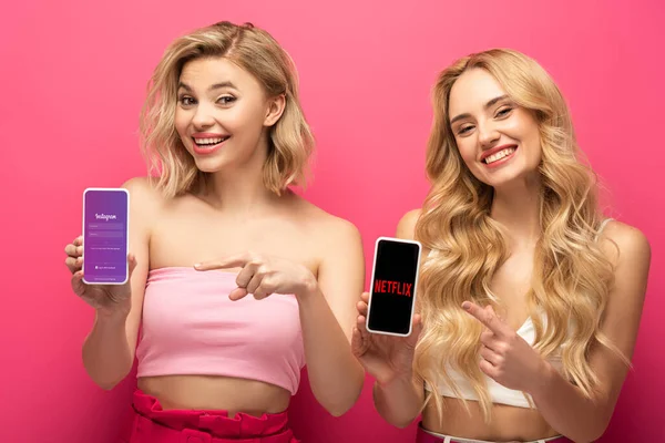 KYIV, UCRANIA - 10 de marzo de 2020: Chicas rubias positivas apuntando a teléfonos inteligentes con aplicaciones de instagram y netflix sobre fondo rosa - foto de stock