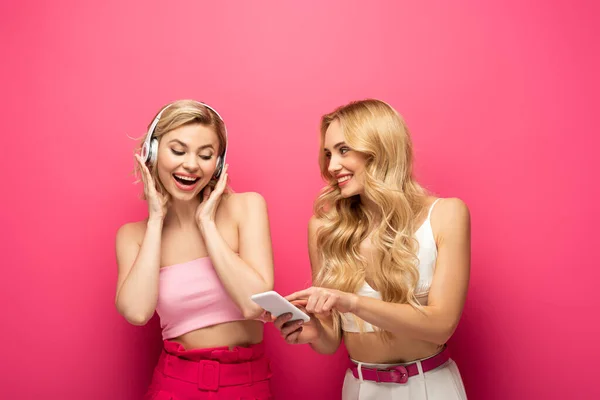 Chica rubia sonriente usando teléfono inteligente cerca de amigo en auriculares sobre fondo rosa - foto de stock