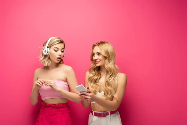 Blonde girl in headphones near smiling friend with smartphone on pink background — Stock Photo