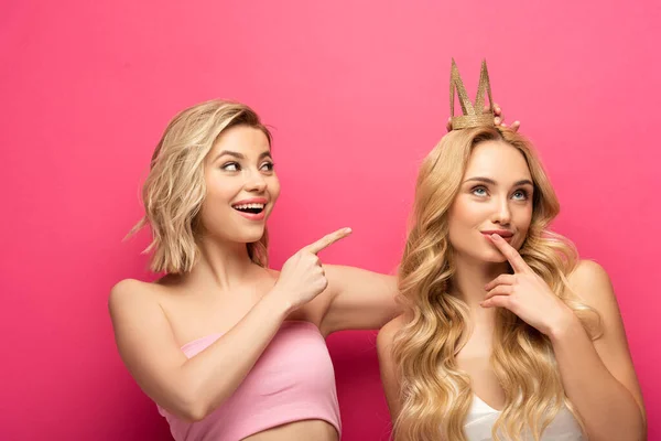 Smiling blonde woman pointing on pensive friend in crown on pink background — Stock Photo