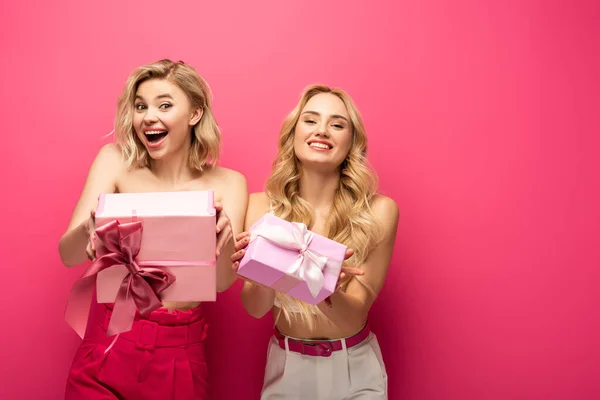 Chicas rubias positivas y conmocionadas sosteniendo cajas de regalo y mirando a la cámara sobre fondo rosa - foto de stock