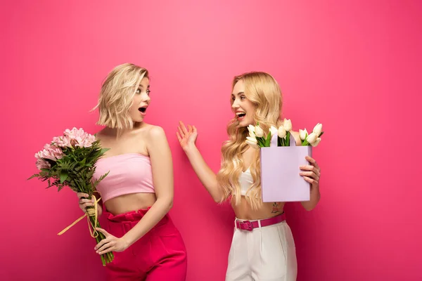 Happy and shocked blonde women looking at each other while holding bouquets on pink background — Stock Photo