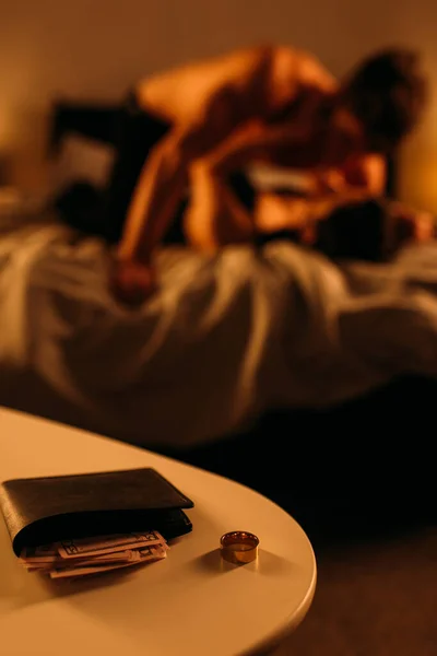 Селективный фокус кошелька с долларовыми банкнотами возле обручального кольца и силуэтами мужчины с проституткой в спальне — стоковое фото