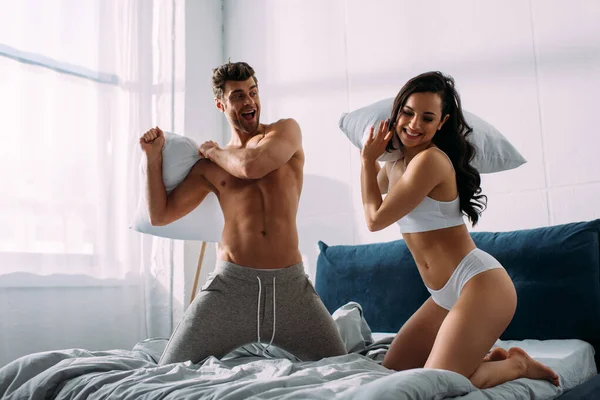 Excited boyfriend and girlfriend smiling and fighting with pillows on bed in bedroom — Stock Photo