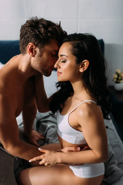 Девушка и парень с закрытыми глазами обнимаются, целуются и сидят на кровати в спальне — стоковое фото