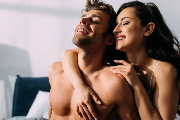 Novia con los ojos cerrados sonriendo y abrazando novio por detrás en el dormitorio - foto de stock