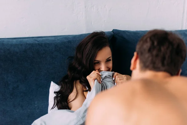 Enfoque selectivo de la mujer cubriendo con manta, sonriendo y mirando al novio en la cama en el dormitorio - foto de stock