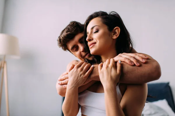 Man with closed eyes smiling and hugging girlfriend from behind in bedroom — Stock Photo