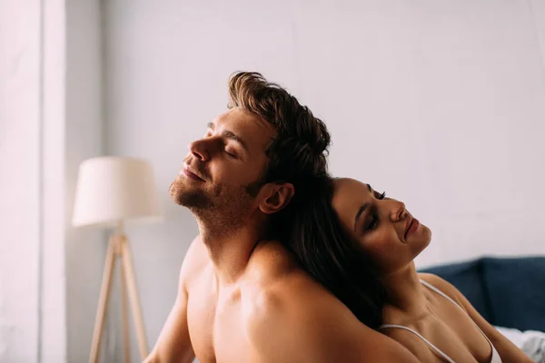 Pareja feliz con los ojos cerrados sentados espalda con espalda en el dormitorio - foto de stock