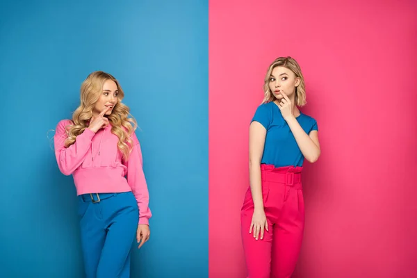 Atractivas hermanas rubias con los dedos cerca de la boca mirando hacia otro lado sobre fondo rosa y azul - foto de stock