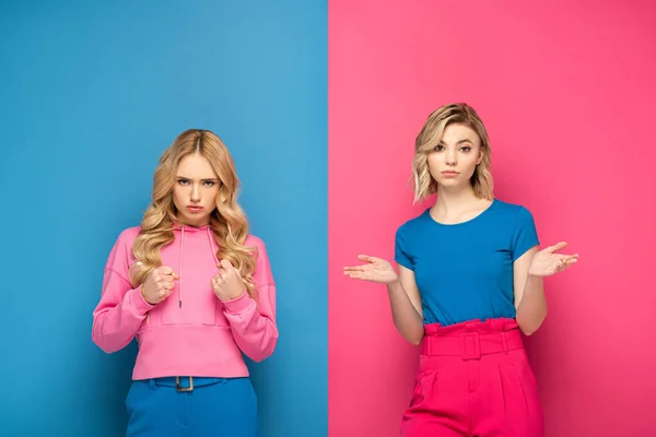 Femme en colère avec les poings regardant la caméra près de soeur blonde montrant signe confus sur fond rose et bleu — Photo de stock