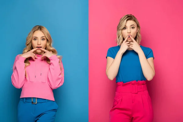 Excited blonde sisters with fingers near lips looking at camera on pink and blue background — Stock Photo