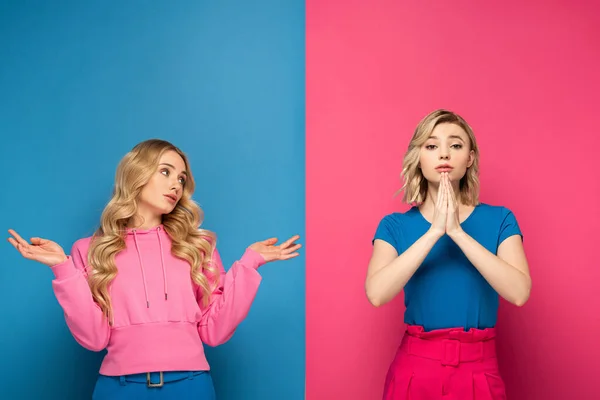Atractiva chica rubia con gesto de encogimiento mirando a la hermana con las manos de oración sobre fondo rosa y azul - foto de stock