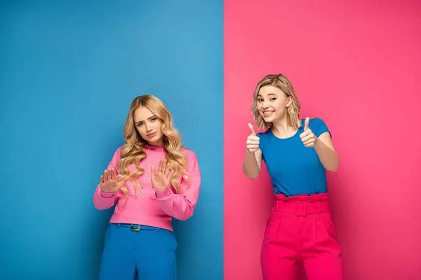 Rubias hermanas mostrando basura y pulgares hacia arriba signos en rosa y azul de fondo - foto de stock