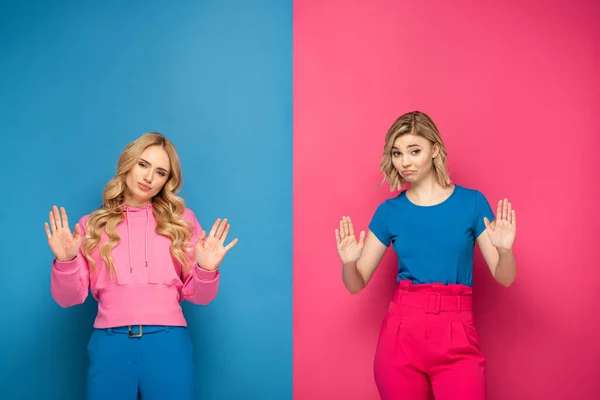 Confused blonde sisters showing stop gesture at camera on pink and blue background — Stock Photo