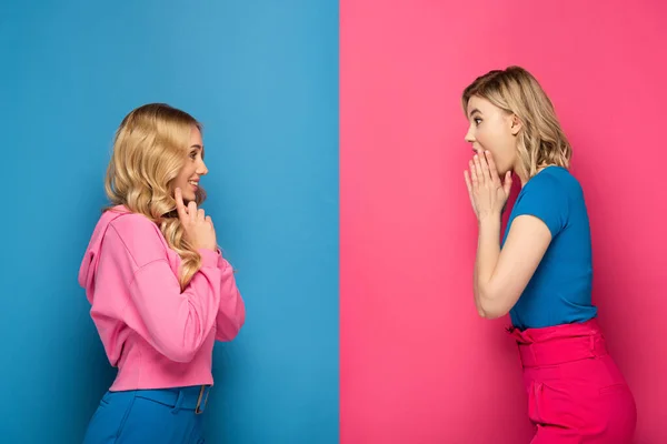Seitenansicht schockierter und lächelnder blonder Schwestern, die einander auf rosa und blauem Hintergrund anschauen — Stockfoto