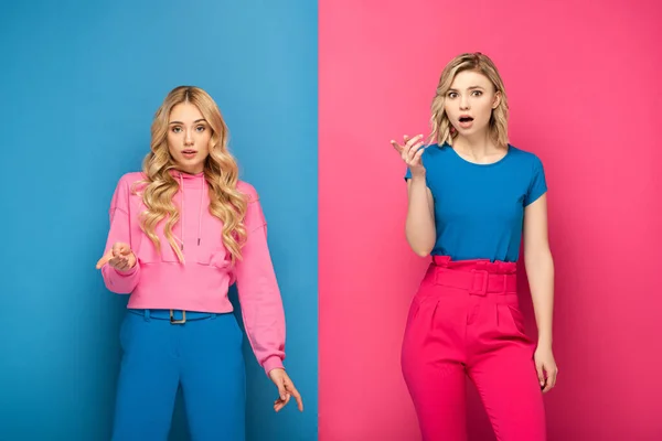 Sœurs blondes excitées pointant avec les doigts sur fond rose et bleu — Photo de stock