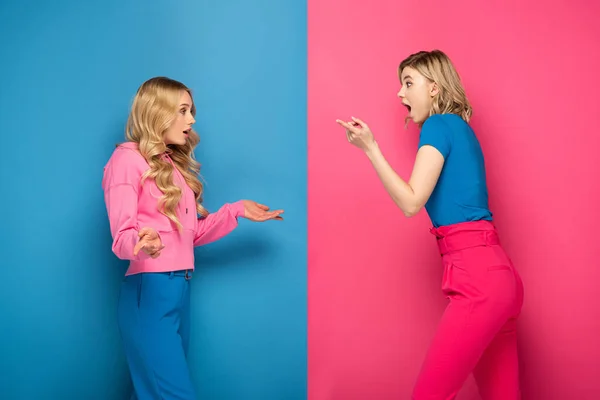 Side view of excited blonde sisters showing shrug gesture and pointing with finger on pink and blue background — Stock Photo