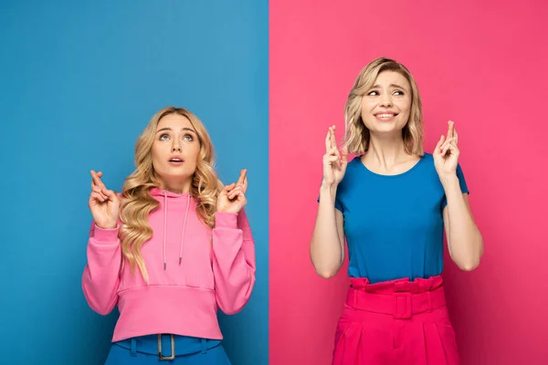 Hermanas rubias con los dedos cruzados mirando hacia otro lado sobre fondo rosa y azul - foto de stock