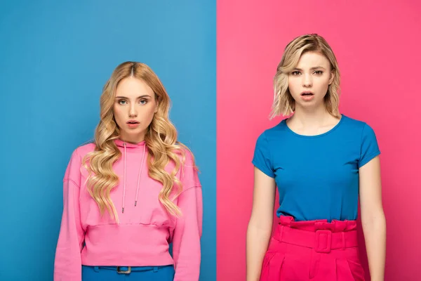 Atractiva chica rubia mirando a la cámara cerca de hermana conmocionada sobre fondo rosa y azul — Stock Photo