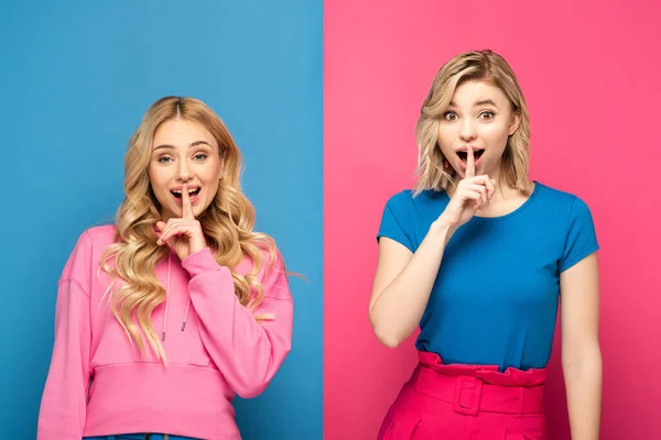 Excited blonde sisters showing silence sign at camera on pink and blue background — Stock Photo