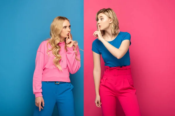 Blonde sisters showing secret sign at each other on pink and blue background — Stock Photo
