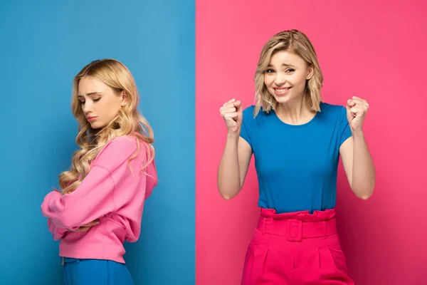Cheerful woman showing yeah gesture near sad blonde sister on pink and blue background — Stock Photo